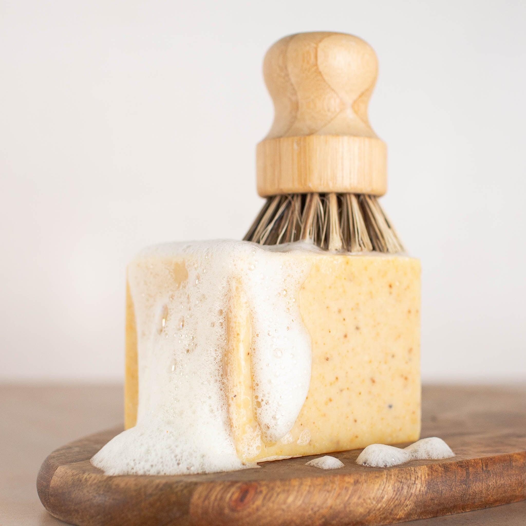 Lathered up pale orange speckled block of vegan Citrus Solid Dish Soap, with dish brush.